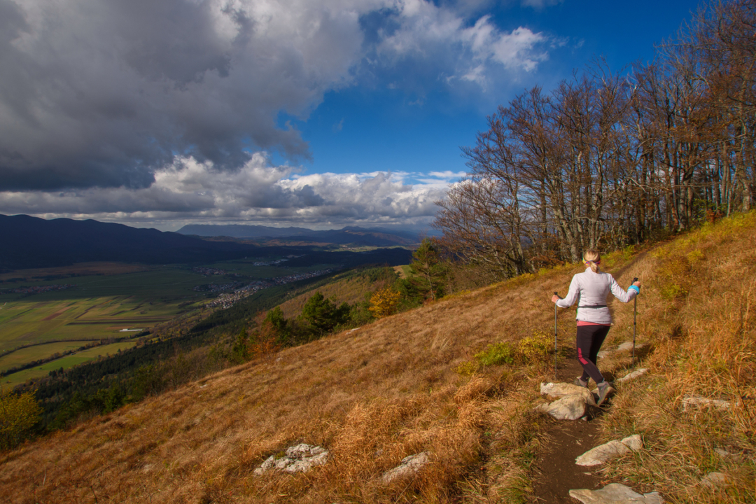 Pohodnica se spušča dol s Slivnice v sončnem jesenskem dnevu. Stopa po ozki stezi čez travnik, odet v rjavkaste jesenske barve. V ozadju je viden prekrasen razgled na okoliške doline ter modro nebo, delno prekrito z oblaki.
