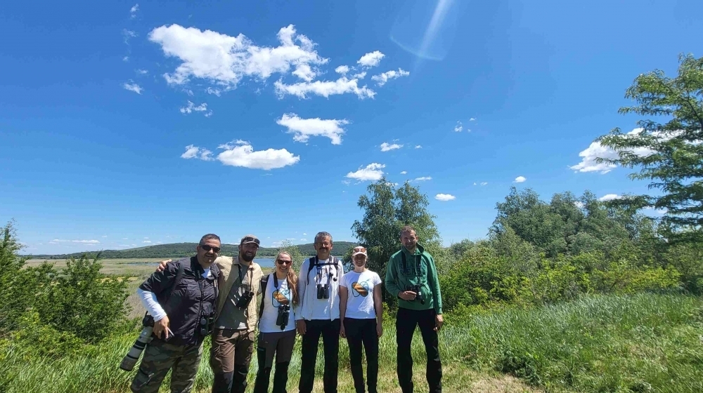 A group of people standing together outdoors in a grassy field on a sunny day, with open landscape and scattered trees in the background under a bright blue sky with a few clouds.