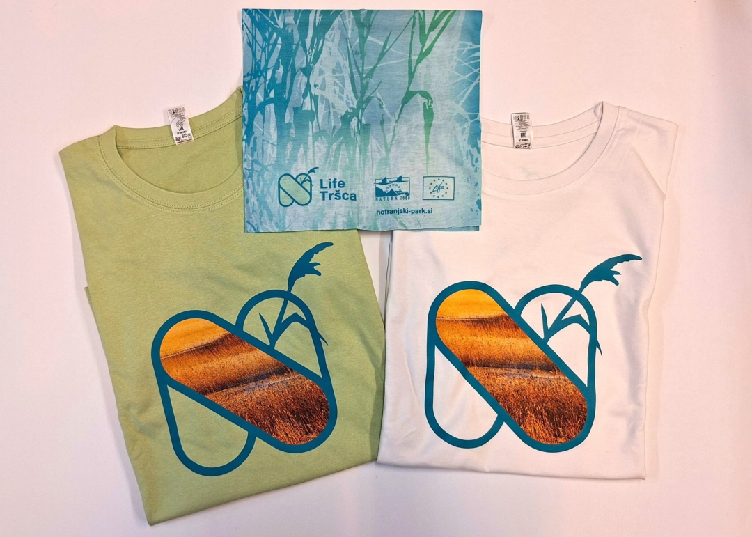 Two folded T‑shirts—one light green and one white—each featuring the Life Tršca logo with a stylized reed design and a photo-textured graphic. Above them lies a blue patterned neck gaiter displaying the Life Tršca logo and partner organization logos.
