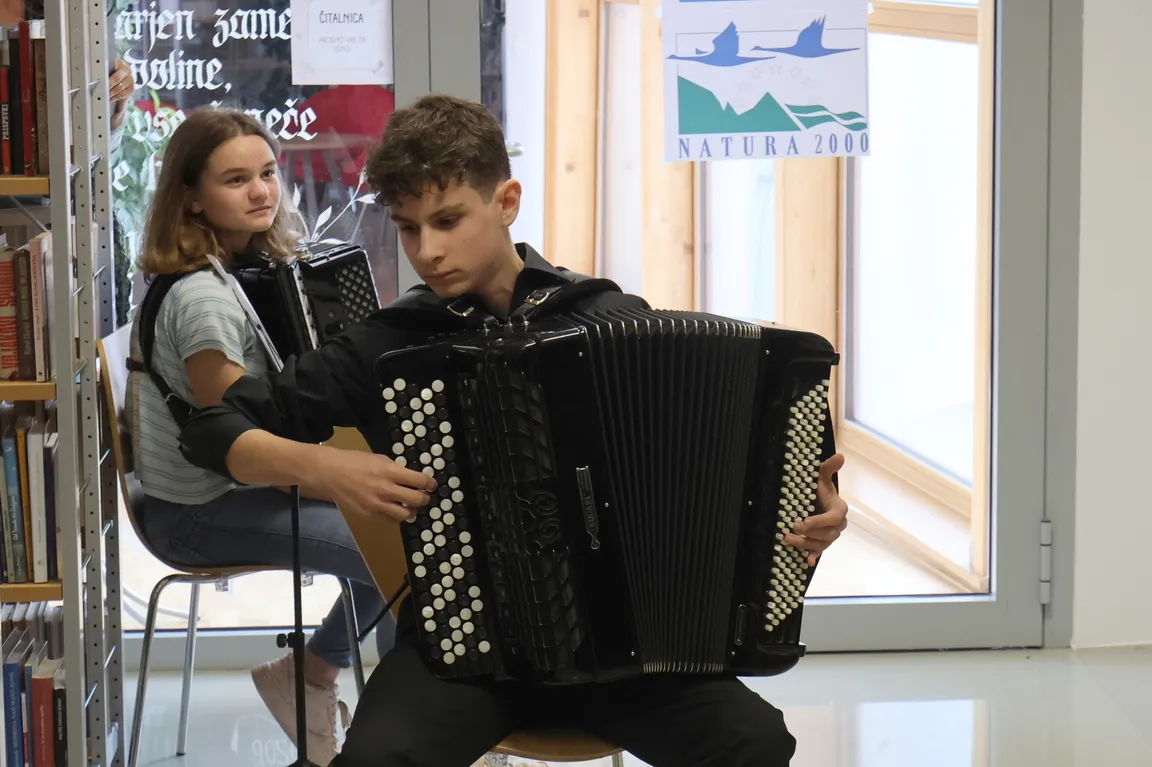 Two people are sitting on chairs; one is playing the accordion. In the background, there is a bookshelf and a window with a poster showing mountains and birds and bearing the text NATURA 2000.