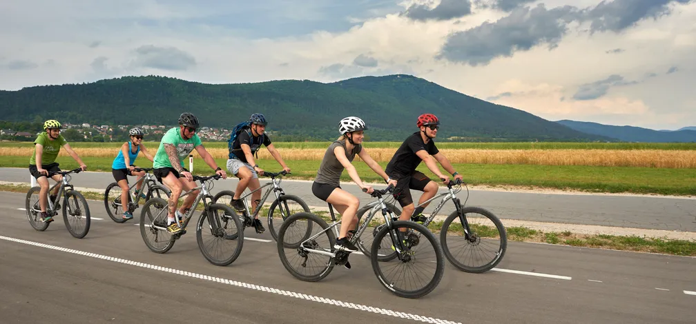 Group of seven cyclists riding along a bicycle road towards Lake Cerknica. In the background fields of grain and mount Slivnica are visible.