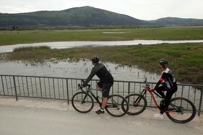Detail from the Witch cycling trail - cyclists standing on a bridge on Lake Cerknica, with mount Slivnica in the background.