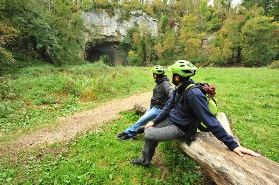 Kolesarja počivata na leseni klopci v Rakovem Škocjanu, v ozadju Veliki naravni most