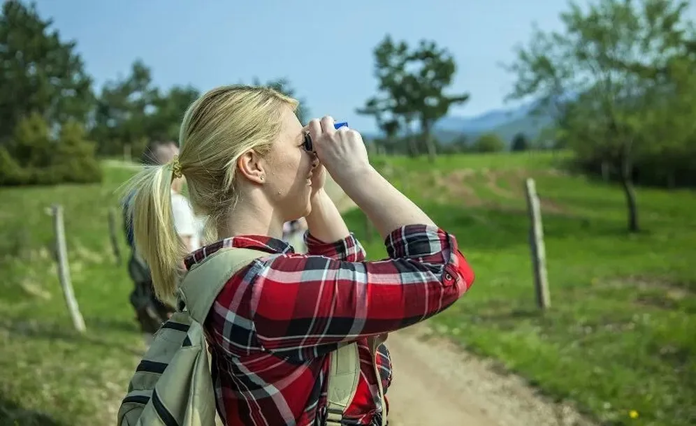 A female visitor in a plaid shirt with blond hair tied in a ponytail watches birds through binoculars. She stands on a gravel path next to a meadow with a couple of trees in the background.