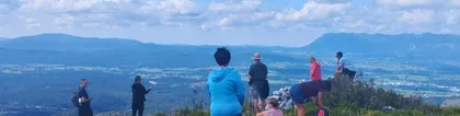 A group of hikers at the Javorniška baba viewpoint overlooking the Pivka Basin, surrounded by forests and hills under a blue sky with clouds.