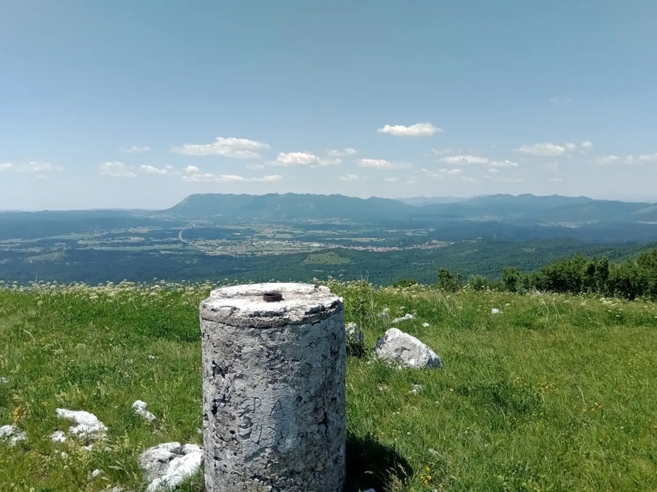 Geodetski kamen stoji na travniku z razgledom na Pivško kotlino in okoliške hribe pod jasnim modrim nebom.