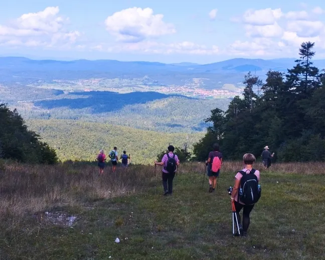 Pohodniki hodijo po travniku na Kaliču z razgledom na Unec, obdanim z gozdovi in hribi pod modrim nebom z oblaki.