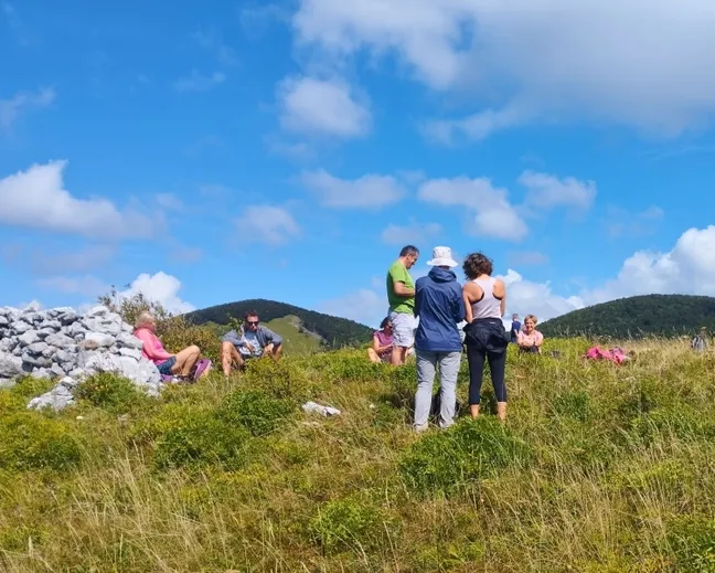 Skupina pohodnikov počiva na travnatem vrhu Javorniške babe z razgledom na okoliške hribe pod modrim nebom z belimi oblaki, ob strani je kup kamenja.