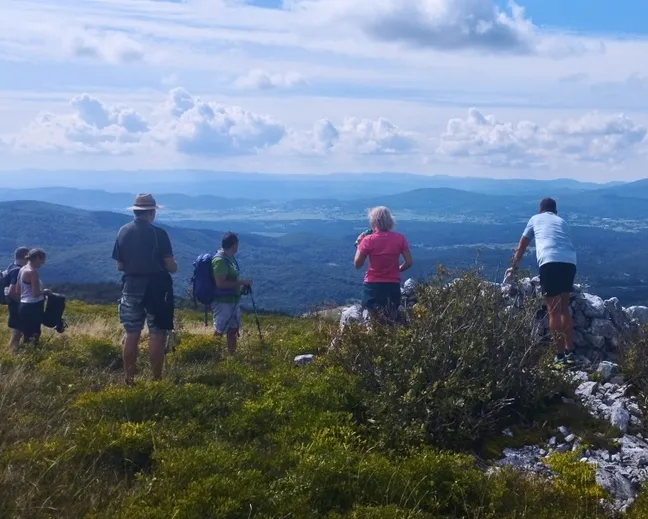 Skupina pohodnikov na razgledni točki Javorniške babe z razgledom na Pivško kotlino, obdano z gozdovi in hribi pod modrim nebom z oblaki.