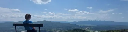 A person sits on a bench at a viewpoint on the summit of Velika Špička and looks out towards the hills and valley under a clear blue sky. The surrounding hills are visible.