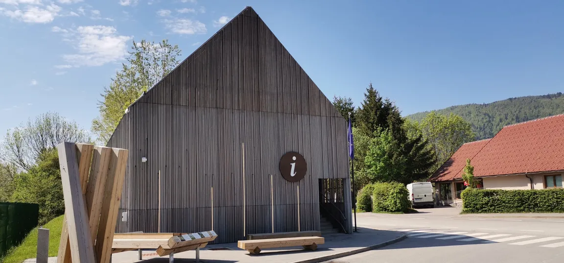 Lesena stavba s strmo, trikotno streho in okroglim znakom z belo črko 'i' na pročelju, ki označuje informacijski center- Center za obiskovalce Cerkniško jezero. V ozadju so drevesa in druge stavbe z rdečimi strehami pod modrim, jasnim nebom.