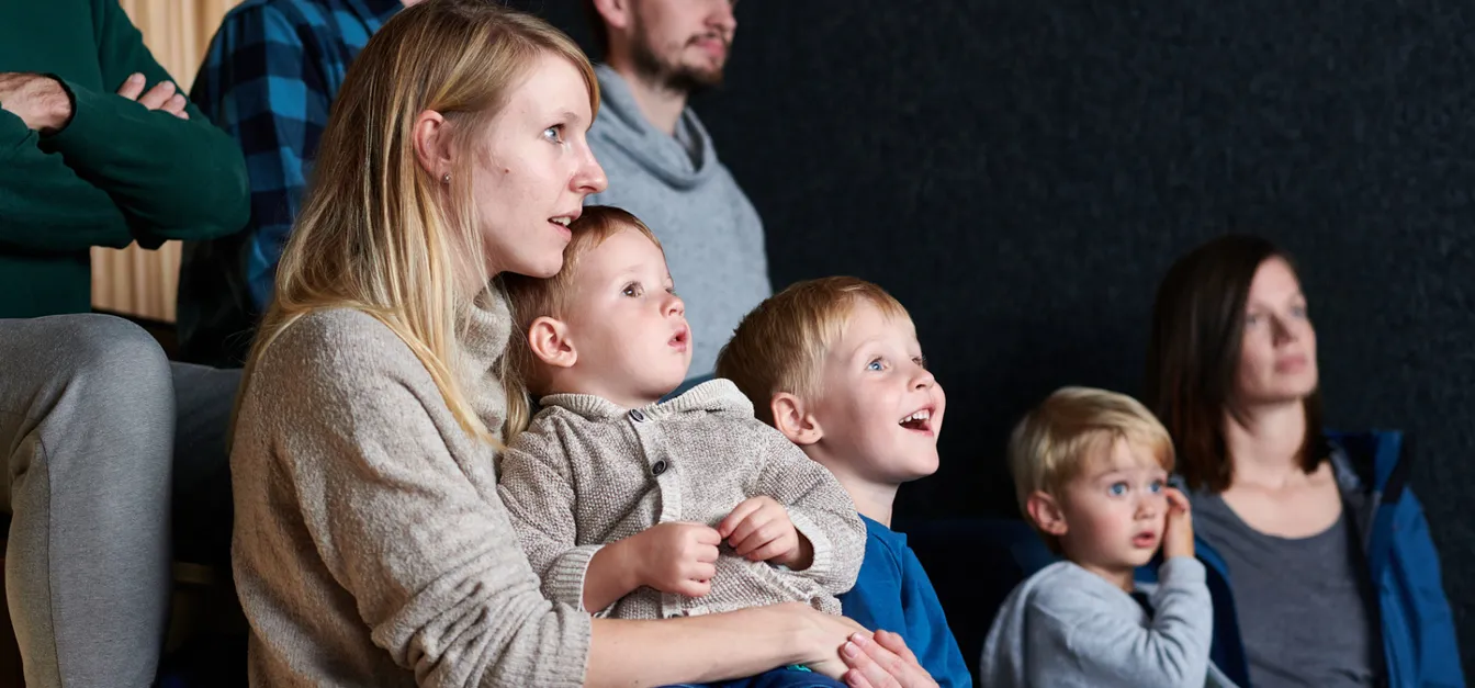 Skupina ljudi, med njimi odrasli in otroci, sedi skupaj in gledajo film v Centru za obiskovalce Cerkniško jezero.