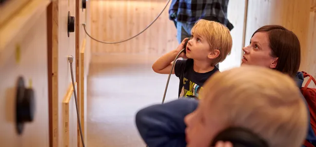 Osebe stojijo pred leseno steno v Centru za obiskovalce Cerkniško jezero, na kateri so pritrjeni črne slušalke. Osebe poslušajo vsebino prek slušalk.