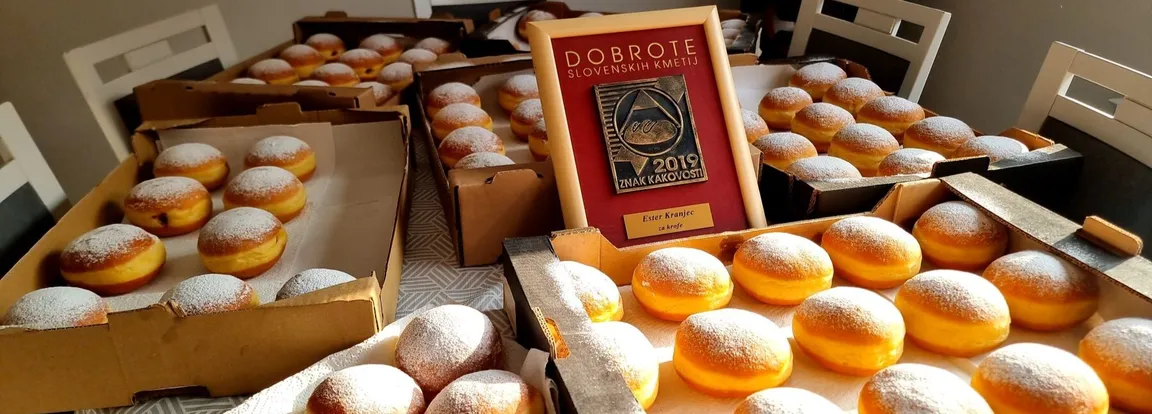 Several trays of freshly baked doughnuts dusted with powdered sugar are displayed on a table, with an award plaque from Dobrote slovenskih kmetij shown among them.