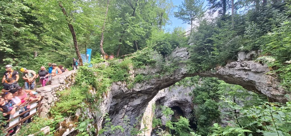 Skupina pohodnikov stoji ob naravnem mostu v Rakovem Škocjanu, ki premošča globoko skalnato dolino, obdano z bujnim zelenjem.