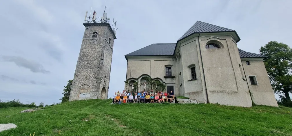 Skupina pohodnikov sedi pred cerkvijo in kamnitim stolpom na vrhu hriba, obdanem z zeleno travo, pod oblačnim nebom.
