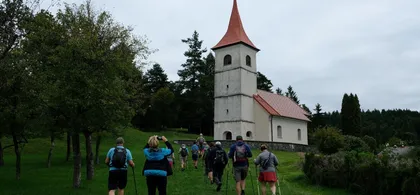 Skupina pohodnikov hodi po travnati poti proti majhni cerkvi z rdečo streho in zvonikom, obdani z drevesi in zeleno pokrajino.