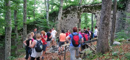 A group of hikers stopping at the ruins of a stone building in the forest, surrounded by tall trees and green foliage.