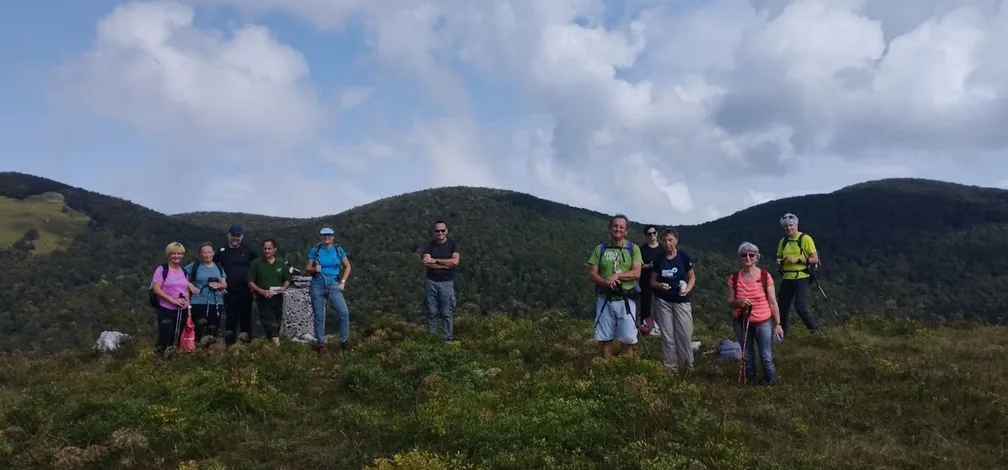 Skupina pohodnikov stoji na travnatem vrhu Javorniške babe, obdana z zelenimi gozdnatimi hribi pod delno oblačnim nebom.