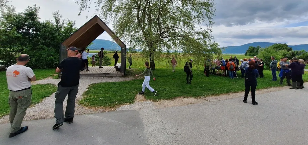 Skupina ljudi se zadržuje ob opazovalnici Malnšče in drevesu ob makadamski poti, v ozadju pa se razprostira zelena pokrajina z gorami pod delno oblačnim nebom.