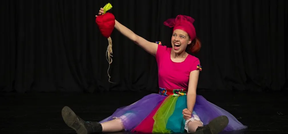 A person is sitting on the floor of a stage with a black background, wearing a colorful skirt in rainbow hues and a red headdress. In one hand, they are holding a red object shaped like a turnip with a long root; their other hand is on the floor. Their black shoes are visible, and their legs are stretched out in front of them.