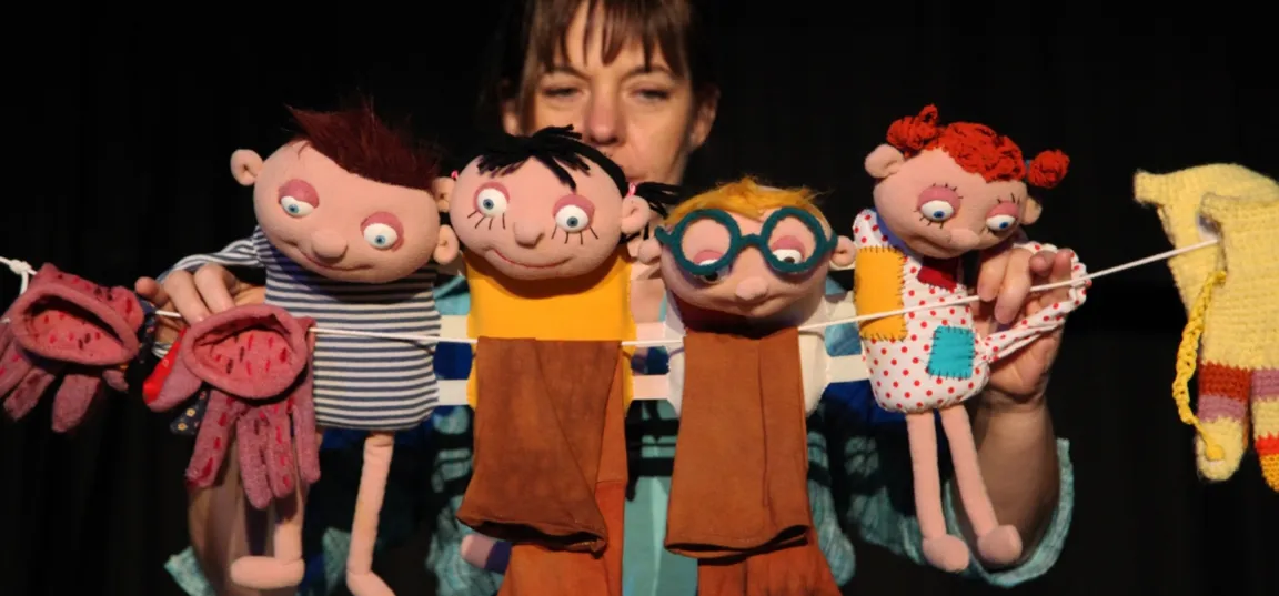 A person behind the puppet stage is holding several hand puppets of various shapes and colors. Among the puppets are figures of children, a puppet with large round glasses, and two smaller animal puppets. The background is dark, so the puppets in the foreground are clearly visible and presented as part of a puppet show.