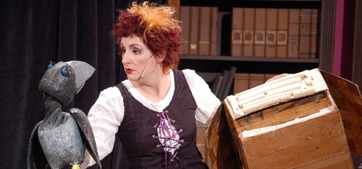 A person is sitting on stage next to a large bird puppet and a wooden suitcase. The person is wearing a dark costume with an embroidered floral pattern, and shelves lined with books are visible in the background. The scene looks like part of a theater or puppet show.