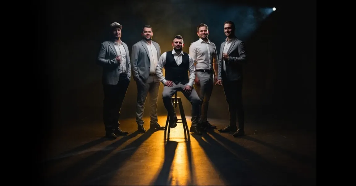 Petčlanska vokalna skupina Il Divji, člani stojijo na zatemnjenem odru, sredinski sedi na barskem stolčku, iz ozadja sveti rumena svetloba.