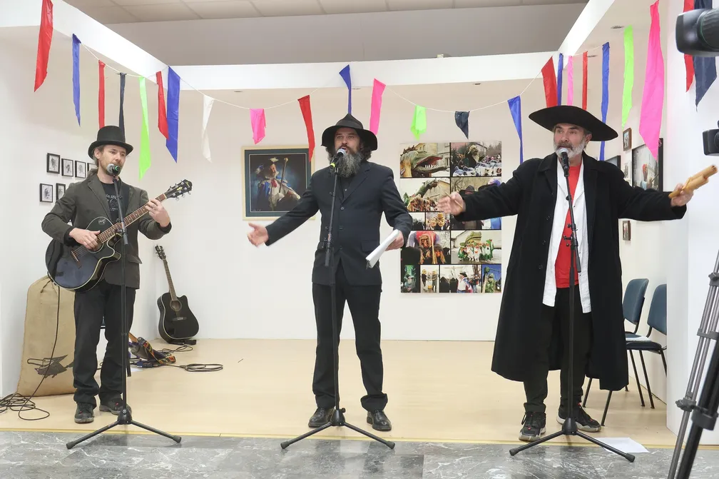 Trije možje v klobukih na mikrofone prepevajo cerkniške pustne pesmi, skrajno levi drži kitaro. Nad njimi visijo okraski ("panklci"), v ozadju pa na steni visijo slike, saj gre za otvoritev pustne razstave.