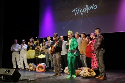 Množica ljudi v maskah stoji na odru za zadnjo točko cerkniške pustne predstave TV Čohovo. Naslov je viden v ozadju na projekciji v Kulturnem domu Cerknica.