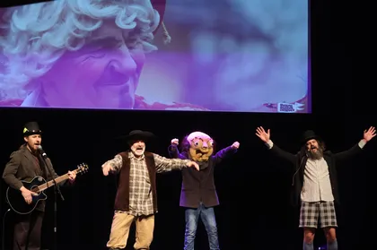 Štirje možje na odru prepevajo in zabavajo navzoče na otroški predstavi v Kulturnem domu Cerknica. Skrajno levi ima v rokah kitaro, dva nosita klobuke, eden pa nosi masko Butalca, tradicionalnega lika cerkniškega pustovanja. V ozadju je na platnu projekcija prizora iz cerkniškega pustnega karnevala.