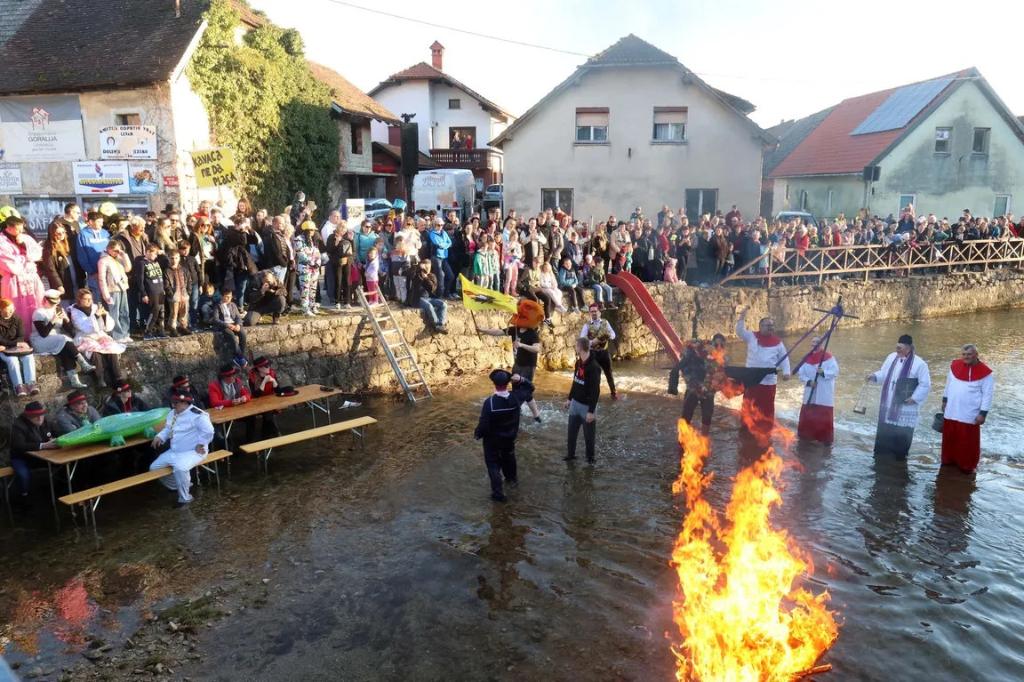 Scene of the burial of "Pust" in Cerknica. Carnival mourners are standing in the Cerkniščica stream, the carnival mascot ("Pust") is already in flames, and a crowd of visitors are watching the events by the stream.