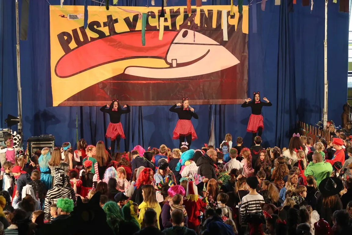 A crowd of children in masks at a children's masquerade in Cerknica, entertained by animators on stage, a banner "Pust v Cerknici" hanging behind them.