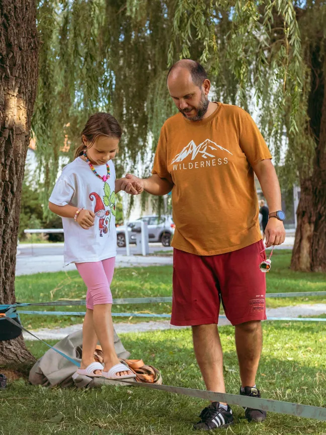 A father helps a girl walk along a slackline stretched between trees.