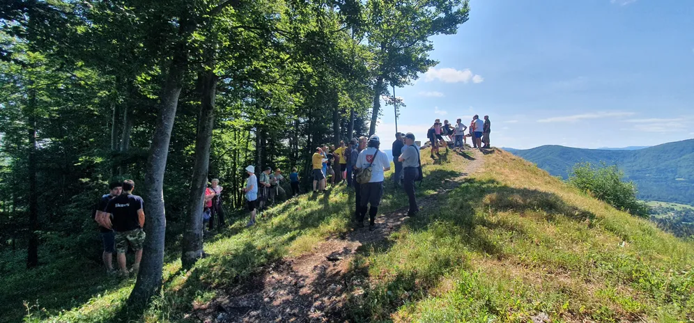 Skupina ljudi je prispela na vrh Špičastega Stražišča pri Selščku. Na levi strani so drevesa, ki nudijo senco, v ozadju pa se razprostirajo hribi pod jasnim sončnim nebom.