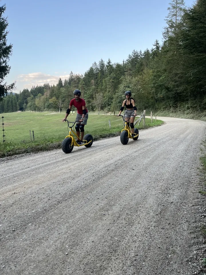 Dve osebi se vozita z gorskimi skiroji po makadamski cesti, obdani z zelenimi polji in drevesi, v ozadju pa je gozd. Nebo je jasno z nekaj oblaki.