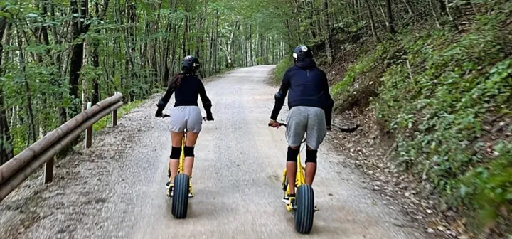 Dve osebi v športnih oblačilih, ščitnikih in čeladah se spuščata po makedamski cesti na dveh rumenih gorskih skirojih. Cesto z obeh strani obdaja narava.