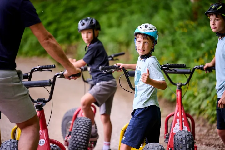 Otroci s čeladami stojijo ob gorskih skirojih na makadamski gozdni cesti, obrnjeni so nazaj.