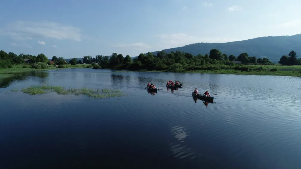 Tri kanuji z več osebami v rdečih rešilnih jopičih plujejo po mirnem jezeru, obdanem z zelenjem in drevesi. V ozadju se razprostira gorska veriga pod jasnim modrim nebom.