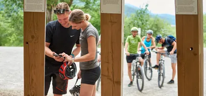 Par na mobilnem telefonu rešuje nalogo, ki je del Kolesarskega doživetja. V ozadju jih čaka skupina treh ljudi na kolesih z zaščitnimi čeladami.
