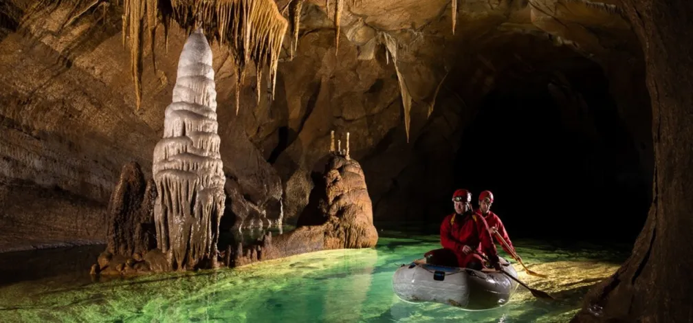 Two people in red clothing and helmets sitting in a small inflatable boat inside a cave. The cave has stalactites hanging from the ceiling and a large stalagmite at the water’s edge. The water is clear with a greenish tint, reflecting the cave formations. The background of the cave is dark, highlighting the illuminated foreground.
