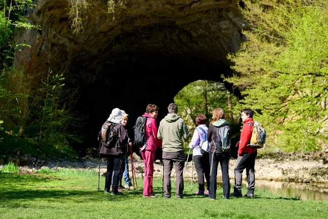 A group of hikers on a guided hike in Rakov Škocjan are gathered on a meadow by the Rak River, with the Big Natural Bridge visible in the background.
