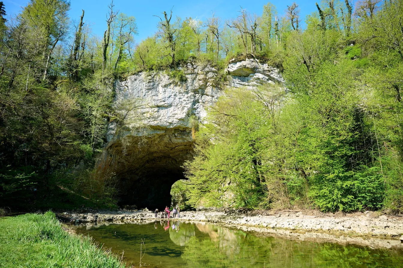 Veliki naravni most v Rakovem Škocjanu, pred njim reka Rak, ob njej pa na kamenju stoji skupina pohodnikov.