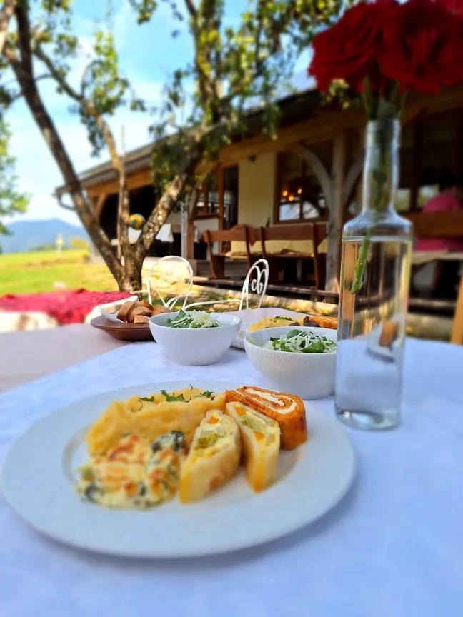 Zunanja miza s krožnikom štrukljev, skledama solate in steklenico vode na sončni terasi.