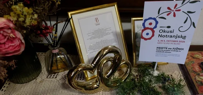 Decorative table at Bajer Estate with a framed Okusi Notranjske 2025 event poster, golden ornament, and floral arrangements.