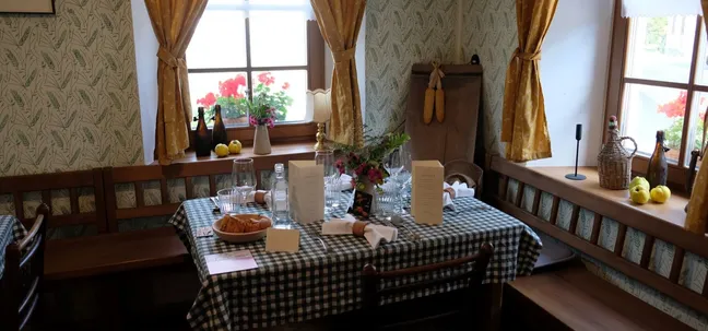 Rustic dining room at Notranjska House – table with checkered cloth, bread, cutlery, flowers, and menus by windows with yellow curtains.