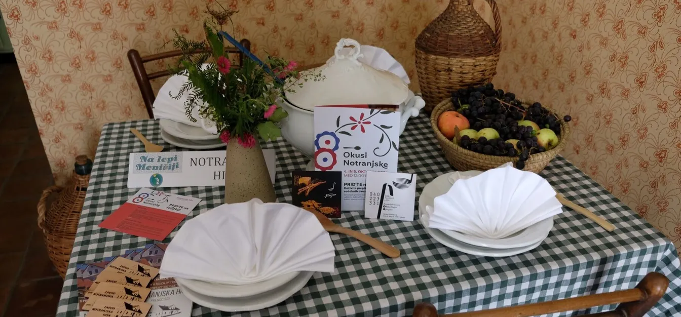 Table setting at Notranjska House – checkered cloth, flowers, basket with grapes and apples, and Okusi Notranjske promotional materials.