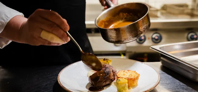 The chef at the Domin Restaurant is preparing the main course, his hands are visible, one holding a pan and the other a spoon with which he pours sauce over a grilled beef steak on a plate.