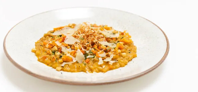 Warm vegetarian appetizer at Gostilnica Domin, creamy risotto with smoked vegetable base, roasted vegetables, goat cheese and Jerusalem artichoke chips on a round white plate on a white background.