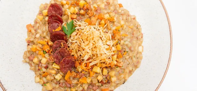 Warm appetizer at Gostilnica Domin, creamy risotto with homemade sausage and root vegetables on a round white plate on a white background, photographed close-up.